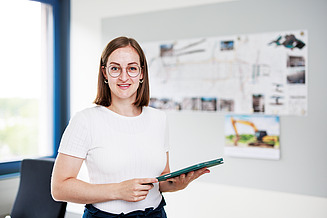 Junge Frau steht mit Tablet im Büro und lächelt in die Kamera.