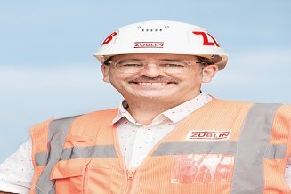 Mitarbeiter mit Schutzhelm und Warnweste auf der Baustelle lächelt in die Kamera. 