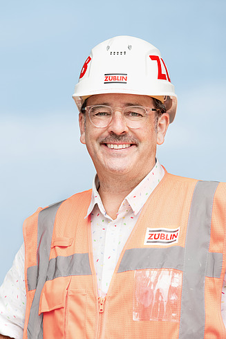 Mitarbeiter mit Schutzhelm und Warnweste auf der Baustelle lächelt in die Kamera. 