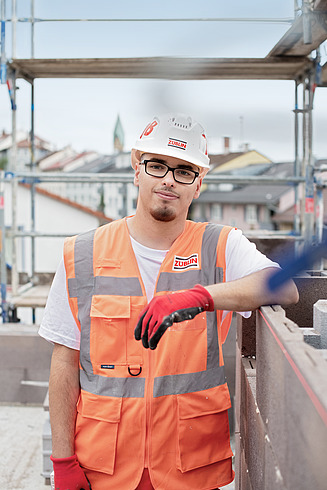 Junger Auszubildender steht mit Warnweste und Schutzhelm auf einer Baustelle.