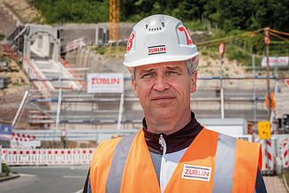 Portrait von einem Mann in Arbeitsschutzkleidung, im Hintergrund ist eine Baustelle zu sehen